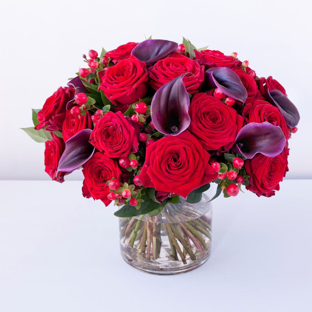 Red roses, wine coloured calla lilies, red hypericum berries in a clear vase