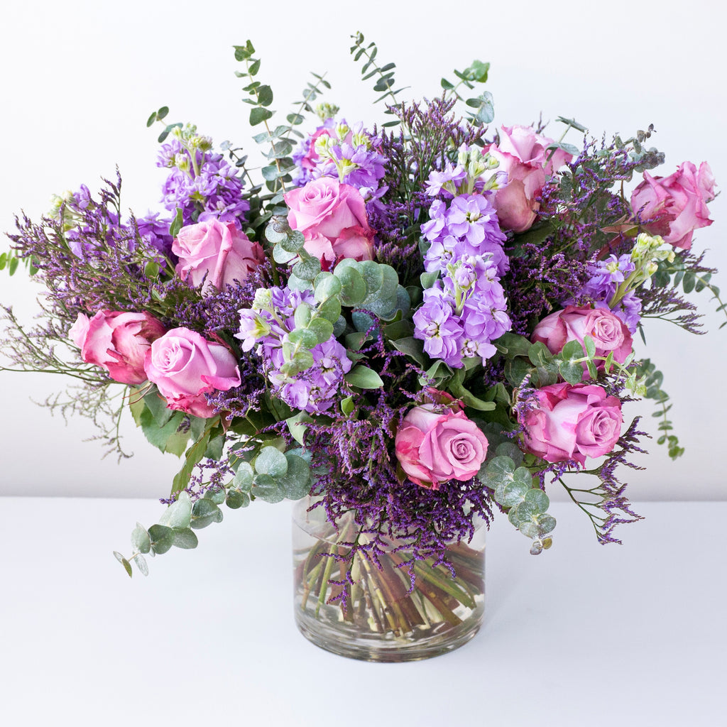 Image of vase of flowers with purple lisianthus, memory lane pink roses, lilac stocks, lilac freesias, with purple statice and foliage.